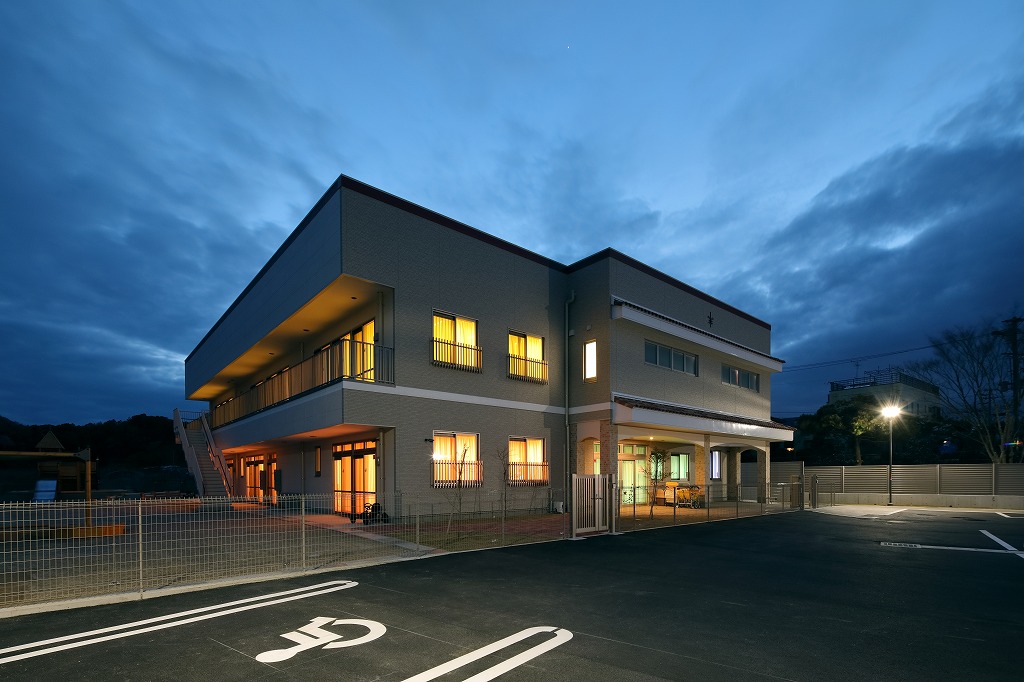 夕暮れ時に電球色のライトが美しく窓から漏れる、真愛三ツ谷こども園の幻想的な外観夜景